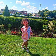 Valentina-Maria participe au concours pour gagner de l'argent avec cette photo : child, sunglasses, heart_shaped, pink_jacket, shorts, sandals, grass, park, hedge, flowers, sunny, blue_sky, clouds, buildings, trees, smiling, outdoor, playful, curly_hair, shadow