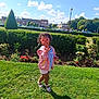 child, sunglasses, heart_shaped, pink_jacket, shorts, sandals, grass, park, hedge, flowers, sunny, blue_sky, clouds, buildings, trees, smiling, outdoor, playful, curly_hair, shadow