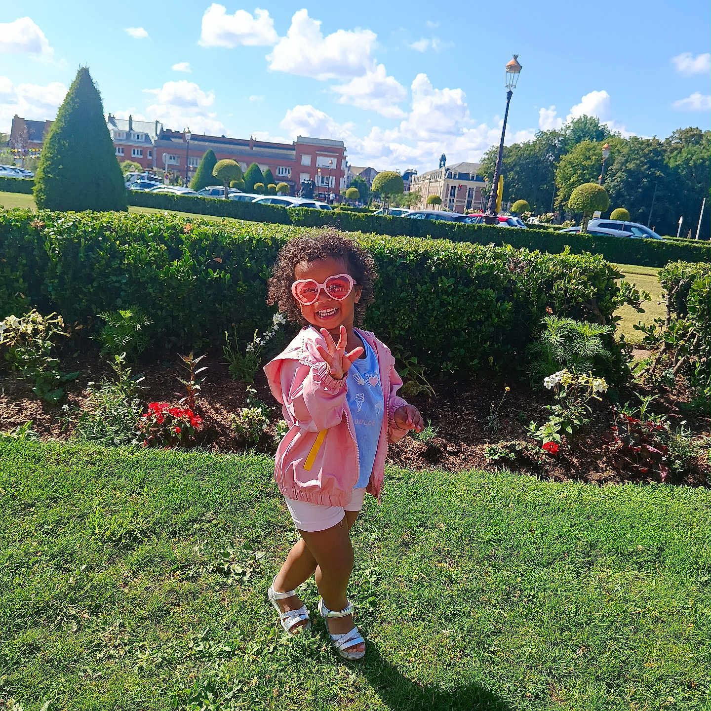 Valentina-Maria participe au concours pour gagner de l'argent avec cette photo : blue_sky, buildings, child, clouds, curly_hair, flowers, grass, heart_shaped, hedge, outdoor, park, pink_jacket, playful, sandals, shadow, shorts, smiling, sunglasses, sunny, trees