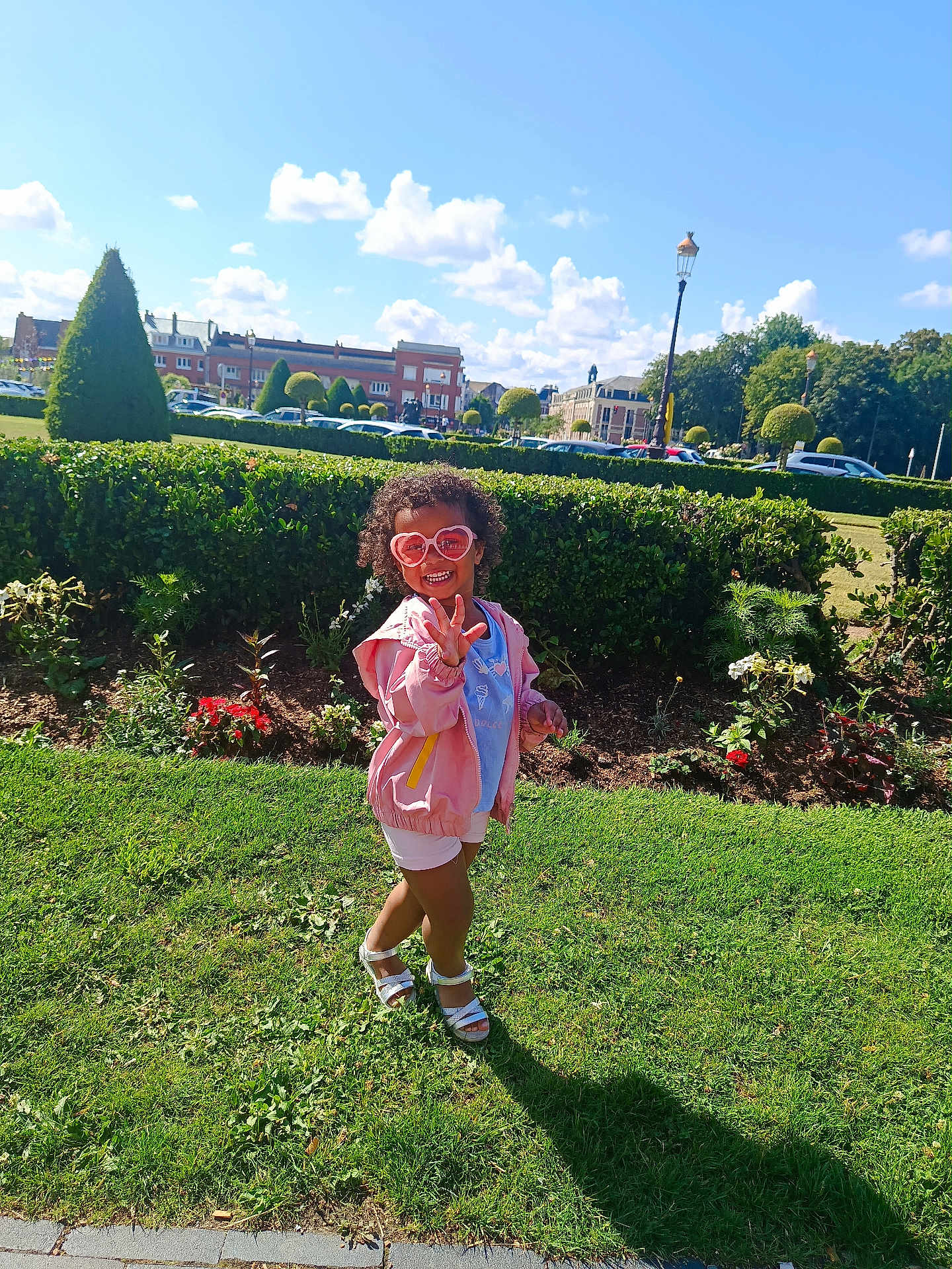 Valentina-Maria participe au concours pour gagner de l'argent avec cette photo : child, sunglasses, heart_shaped, pink_jacket, shorts, sandals, grass, park, hedge, flowers, sunny, blue_sky, clouds, buildings, trees, smiling, outdoor, playful, curly_hair, shadow