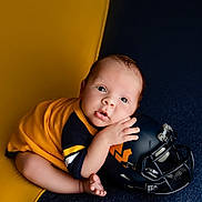 Caden joined the competition — help win amazing prizes! baby, infant, child, football, helmet, sports, portrait, indoors, sitting, lying_down, smiling, wide_eyes, hands, bare_feet, yellow, navy_blue, clothing, photography, close_up, adorable