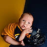 baby, infant, child, football, helmet, sports, portrait, indoors, sitting, lying_down, smiling, wide_eyes, hands, bare_feet, yellow, navy_blue, clothing, photography, close_up, adorable