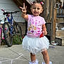 bicycles, birthday_girl, chalk_drawing, child, concrete, curly_hair, daylight, frilly_socks, hair_bows, happy, holding_snack, outdoor, peace_sign, person, pink_sneakers, pink_tshirt, playful, smiling, stairs, white_tutu