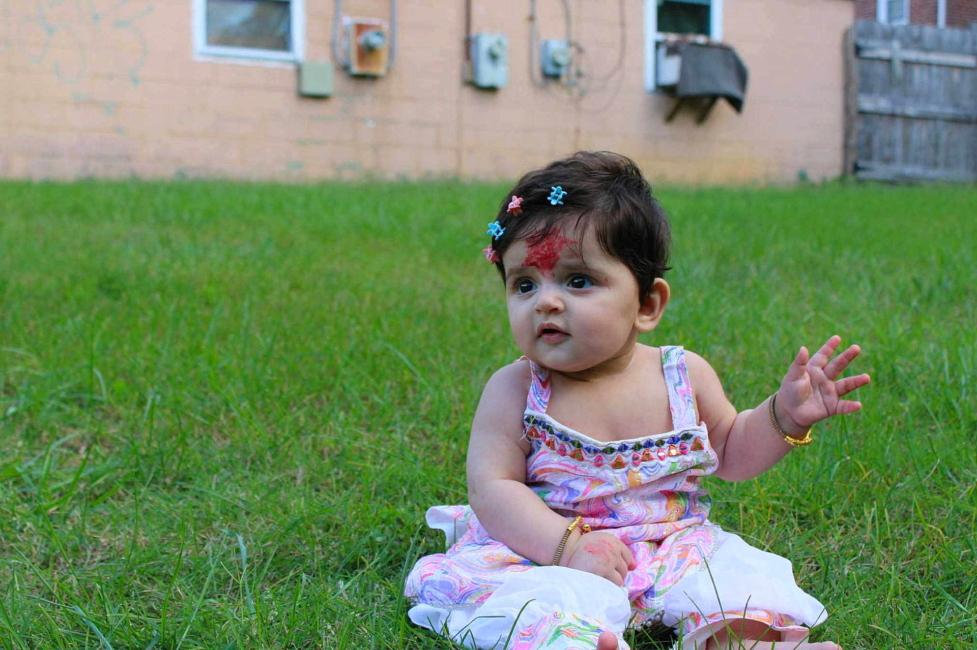 Nayasha is registered to the contest to win money with this photo: baby, background, bracelet, child, curious, cute, daylight, dress, forehead_mark, garden, grass, greenery, hair_clips, infant, nature, outdoor, person, sitting, skin, young