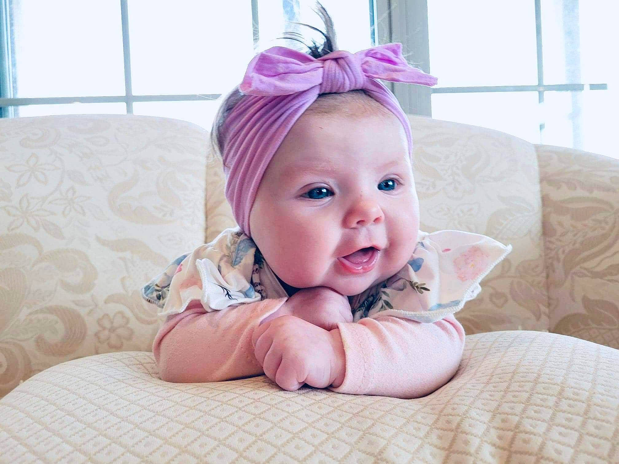 Delilah is registered to the contest to win money with this photo: baby, baby_products, baby_toddler_clothing, cheek, comfort, eye, fashion_accessory, fun, grass, hair_accessory, happy, headband, headpiece, linens, magenta, person, pink, purple, room, smile