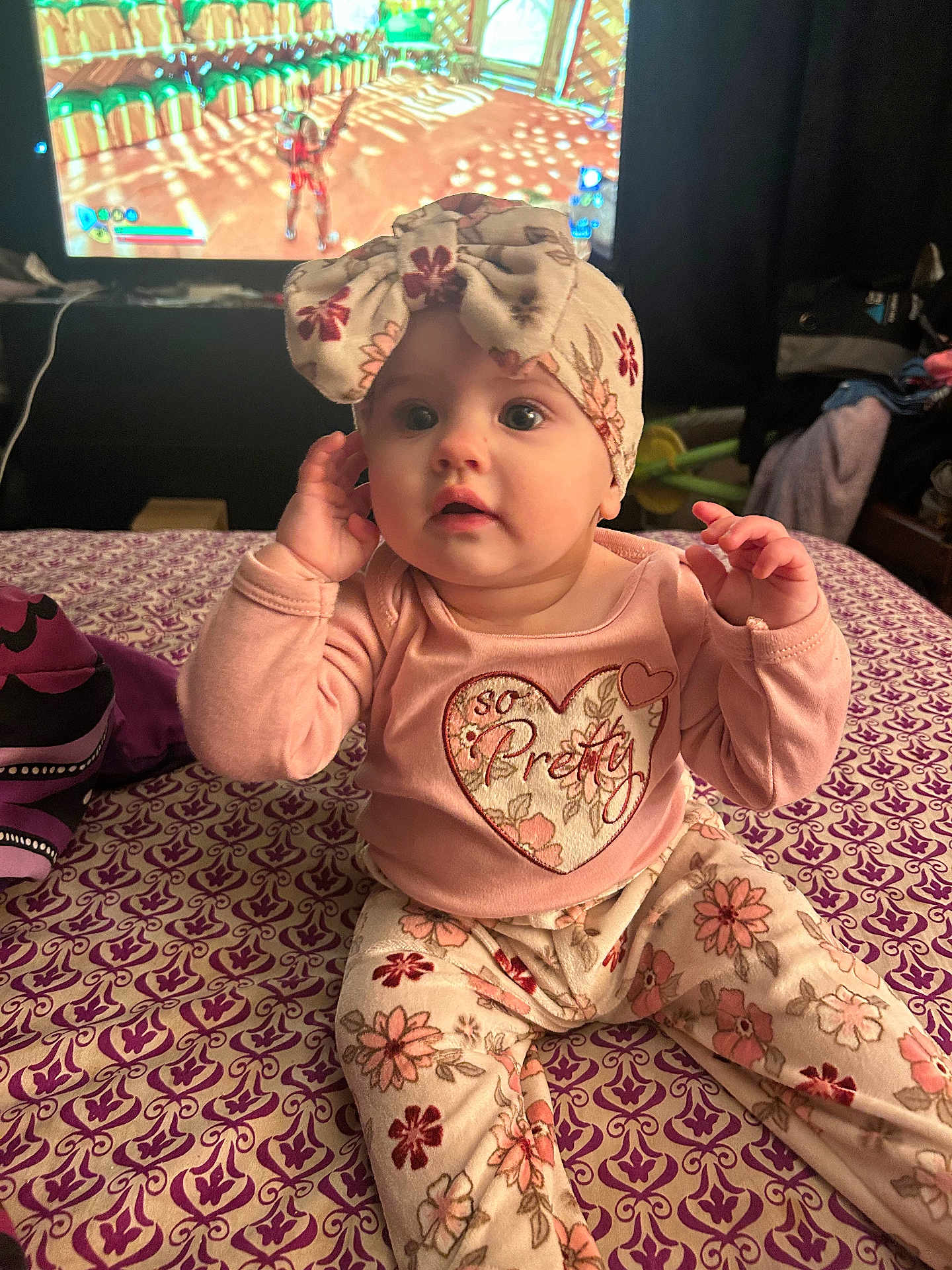 Kyrah is registered to the contest to win money with this photo: baby, child, floral_bow, floral_pants, pink_shirt, patterned_surface, indoor, television, video_game, curious, sitting, clothing, person, headwear, cute, home, living_room, screen, play, young