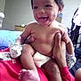 baby, infant, smile, dimples, mittens, diaper, hand, person, indoor, bed, furniture, shirt, red_clothing, skin, foot, happy, child, room, adult, holding