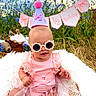 Elena is registered to the contest to win money with this photo: baby, child, sunglasses, party_hat, pink_dress, flowers, outdoor, grass, plush_toys, banner, celebration, nature, portrait, cute, smiling, sunlight, grass_field, fashion, happy, infant