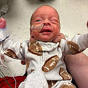 Carter is registered to the contest to win money with this photo: newborn, baby, infant, hospital, nasal_tube, medical_tape, onesie, football_pattern, person, winking, holding, cute, small, human, child, bedroom_furniture, floor, healthcare, caregiver_hand, clothing