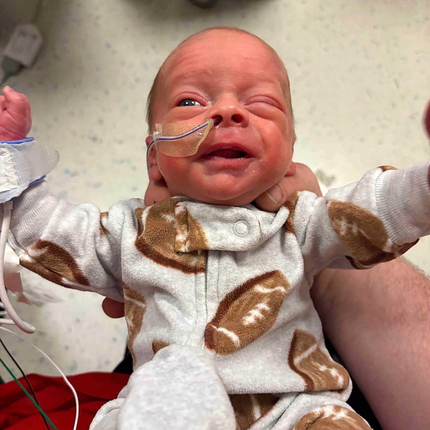 Carter is registered to the contest to win money with this photo: newborn, baby, infant, hospital, nasal_tube, medical_tape, onesie, football_pattern, person, winking, holding, cute, small, human, child, bedroom_furniture, floor, healthcare, caregiver_hand, clothing
