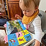 Sébastien participe au concours pour gagner de l'argent avec cette photo : toddler, child, high_chair, book, reading, bib, indoor, wooden_furniture, table, floor, person, baby, curious, colorful, picture_book, home, decor, seated, hand, clothing