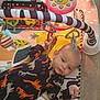 baby, infant, play_mat, toy, dinosaur_print, onesie, colorful, hanging_toys, floor, indoors, child, cute, soft_lighting, face, hand, head, pattern, playtime, baby_clothes, expression