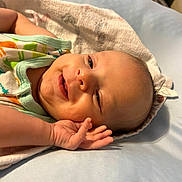 Enzo is registered to the contest to win money with this photo: baby, smiling, infant, hand, face, blanket, onesie, dinosaur_print, lying_down, soft_surface, cute, child, newborn, skin, head, expression, cozy, relaxed, indoors, portrait