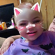 Peyton joined the competition — help win amazing prizes! child, toddler, purple_shirt, halloween, bat, ghost, glitter, face_filter, cat_ears, whiskers, indoor, wood_paneling, smile, happy, person, casual, seated, family, playful, digital_filter