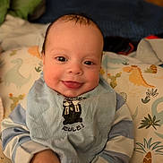 Quentin is registered to the contest to win money with this photo: baby, infant, smiling, tongue_out, pillow, dinosaur_pattern, bib, striped_pajamas, cute, face, child, indoors, happy, lying_down, young_child, person, soft_lighting, closeup, cozy, background