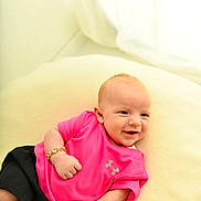 Ronnie Trevor joined the competition — help win amazing prizes! baby, infant, smile, pink_shirt, black_shorts, bracelet, cushion, indoor, happy, cute, child, person, portrait, soft_lighting, relaxed, clothing, skin, face, small_hands, lying_down
