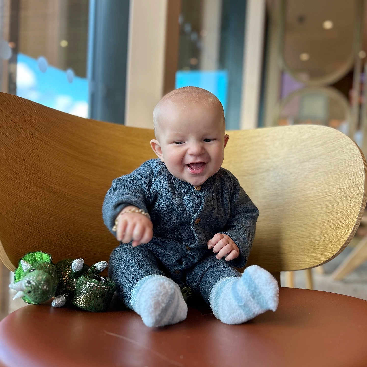Ronnie Trevor is registered to the contest to win money with this photo: baby, blurred_background, chair, child, cozy, cute, dinosaur_toy, gray_outfit, happy, indoor, infant, joyful, person, plush_toy, portrait, sitting, smiling, socks, window, wooden_chair