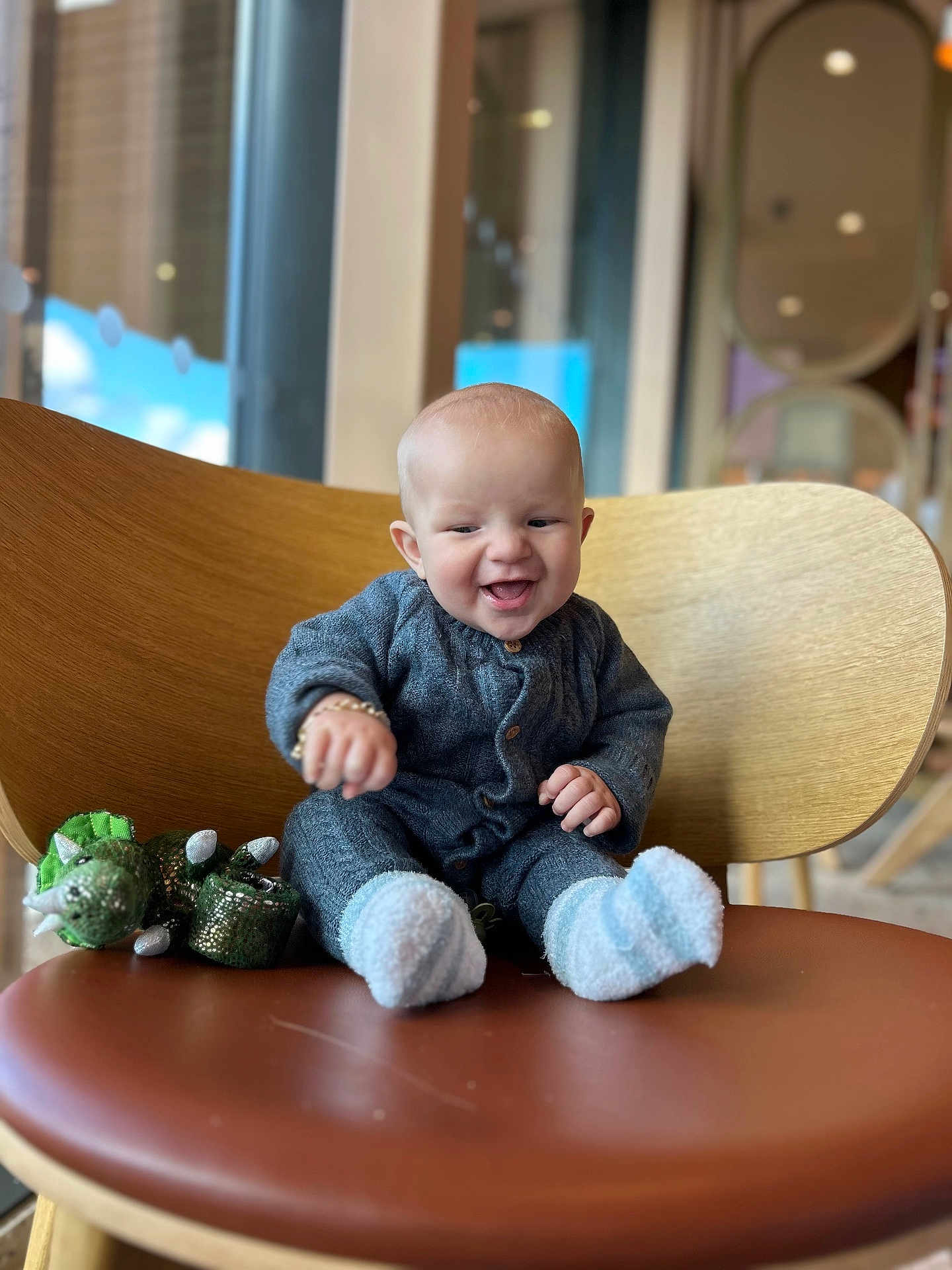 Ronnie Trevor is registered to the contest to win money with this photo: baby, child, smiling, happy, sitting, chair, wooden_chair, plush_toy, dinosaur_toy, gray_outfit, socks, indoor, window, blurred_background, cute, infant, person, portrait, cozy, joyful