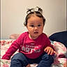 baby, child, infant, sitting, bed, bedroom, floral_bedding, toy, green_toy, long_sleeves, pants, bare_feet, hair_bows, curious, indoors, portrait, face, smiling, furniture, social_media_screenshot