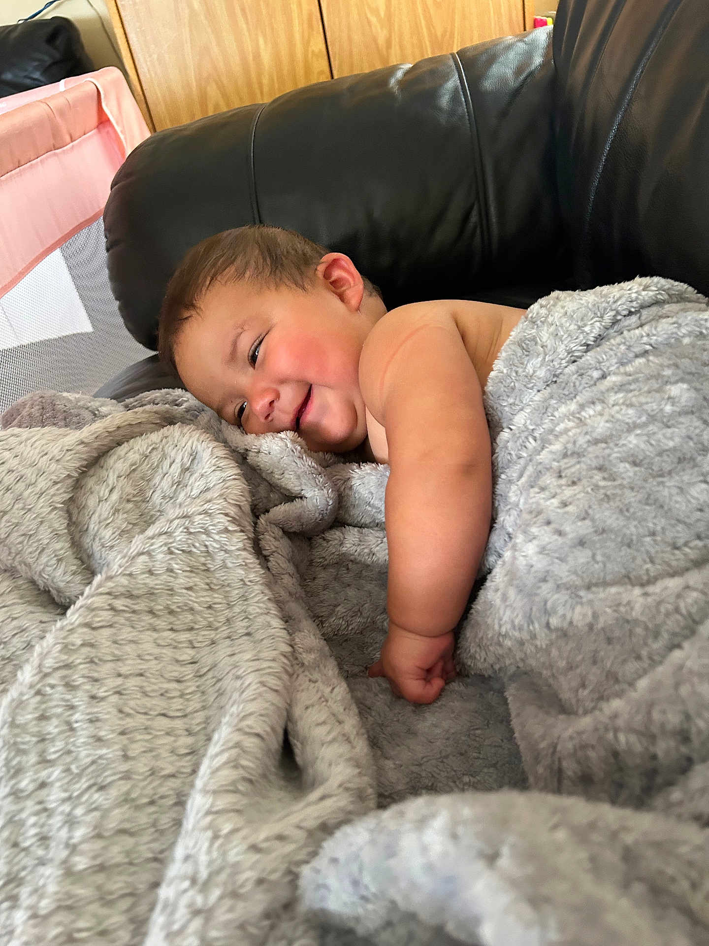 Charlee is registered to the contest to win money with this photo: baby, smiling, blanket, couch, black_leather, infant, wooden_cabinet, happy, cozy, indoor, child, face, skin, arm, furniture, relaxing, comfort, home, portrait, expression