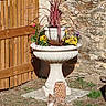 dog, puppy, small_dog, long_hair, brown_fur, white_fur, lying_down, outdoor, garden, flowers, flower_pot, planter, urn, stone_wall, wooden_gate, rustic, lantern, patio, grass, sunlight