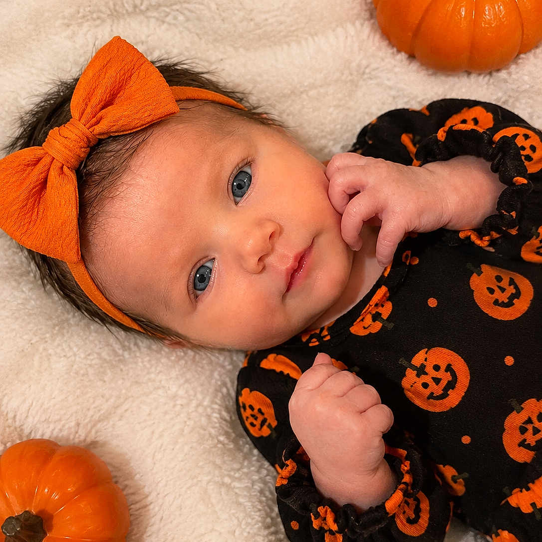 Ashlynn is registered to the contest to win money with this photo: baby, infant, child, portrait, halloween, pumpkin, bow, headband, onesie, orange, black, blanket, lying_down, indoors, close_up, face, eyes, smiling, cozy, festive