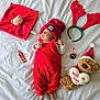 baby, red_overalls, red_hat, teddy_bear, christmas, holiday, bed, white_bed_sheet, reindeer_antlers, pine_cone, plush_toy, infant, cute, festive, decorations, lying_down, soft_toy, christmas_hat, child, indoors
