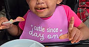 Jasmine is registered to the contest to win money with this photo: toddler, child, high_chair, bib, food, spoon, plate, curly_hair, smiling, meal, indoor, table, smartphone, cartoon, television, animation, pink, happy, face, person