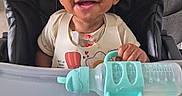 Titus is registered to the contest to win money with this photo: baby, child, high_chair, smiling, bottle, strawberry, food, feeding, indoor, person, cute, happy, infant, table, seat, plastic, clothing, curly_hair, face, young
