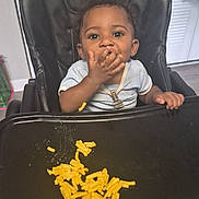 Titus is registered to the contest to win money with this photo: baby, child, high_chair, food, macaroni, cheese, messy, indoor, person, feeding, clothing, face, hand, tray, seat, black, happy, cute, young, infant