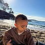 toddler, child, beach, sea, water, sky, sunlight, pebbles, jacket, brown_clothing, outdoor, nature, daylight, portrait, young_child, coast, calm, focused, casual, scenic