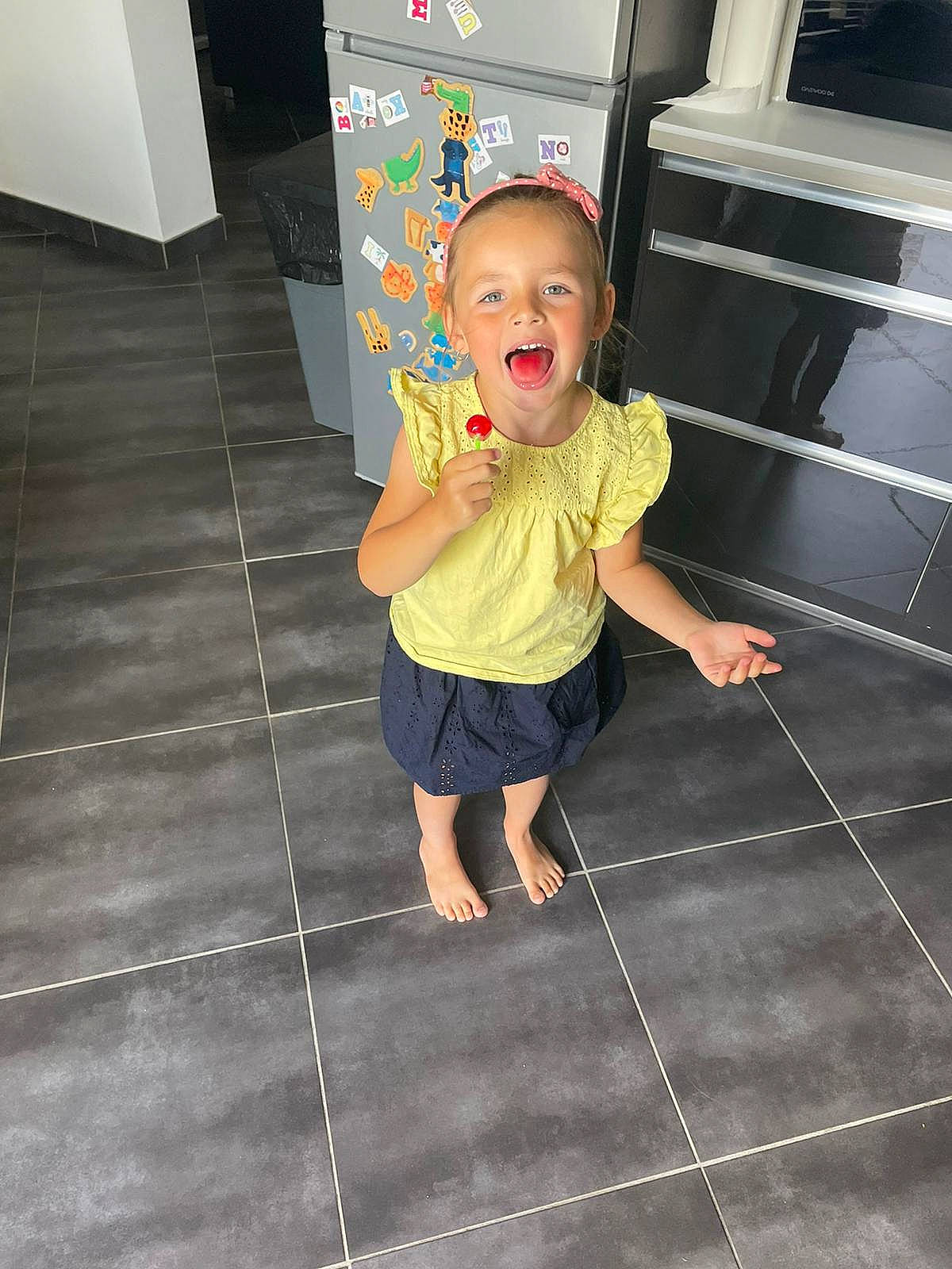 Nina a rejoint le concours — aidez-le/la à gagner de superbes lots ! baby_toddler_clothing, barefoot, black, child, concrete, floor, flooring, foot, fun, hardwood, human_leg, leg, pattern, person, play, road_surface, sandal, t_shirt, tile, toddler