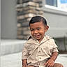 background_blur, beige_shirt, brown_pants, casual_clothing, child, concrete, cute, daylight, face, hair, happy, outdoor, person, portrait, short_sleeves, sitting, smiling, stone_wall, toddler, window