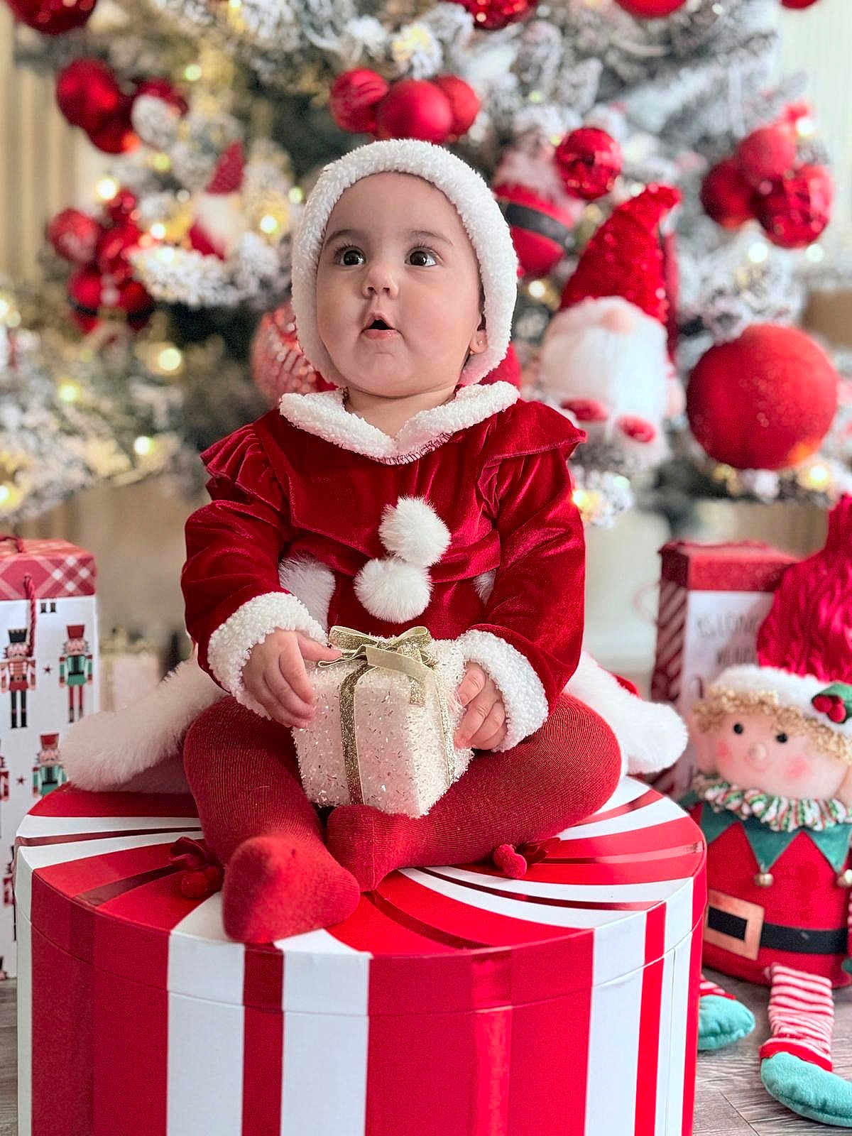 Camelia is registered to the contest to win money with this photo: baby, child, christmas, holiday, gift, present, red_clothing, hat, festive, decorations, tree, ornaments, toy, doll, floor, indoor, cute, smiling, sitting, seasonal