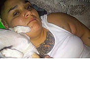 Peewee is registered to the contest to win money with this photo: person, dog, white_dog, pet, paw, face, bed, bedroom, blanket, pillow, lying_down, indoor, portrait, tattoo, white_tank_top, relaxed, funny, companionship, cuddling, flash_photography