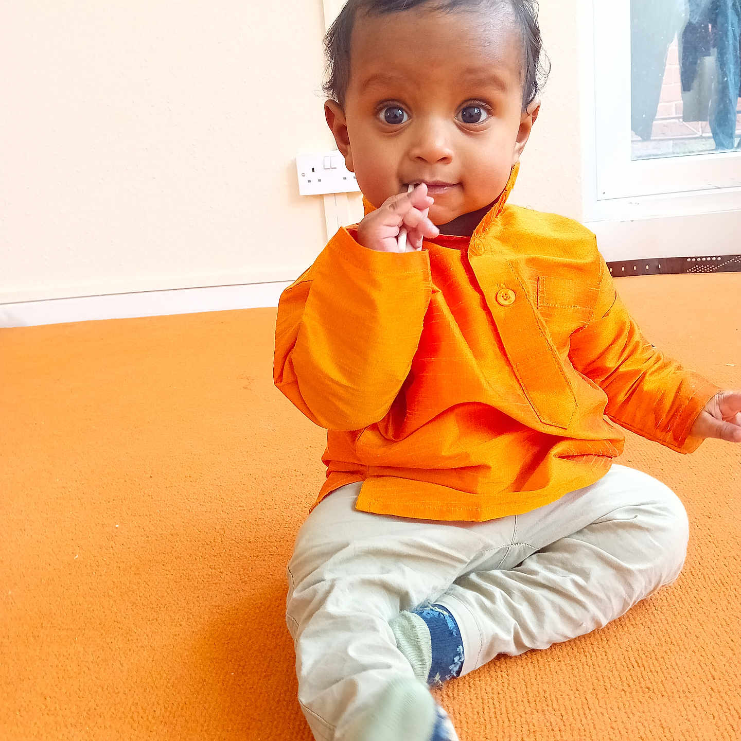 Adheesh is registered to the contest to win money with this photo: baby, carpet, child, clothing, curly_hair, cute, expression, face, floor, hand, indoor, light, orange_shirt, pants, person, portrait, sitting, sock, window, young