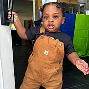 Yosiah joined the competition — help win amazing prizes! toddler, child, overalls, black_shirt, door_handle, indoor, kitchen, curly_hair, pigtails, young_child, standing, expression, person, floor, cabinet, refrigerator, bag, colorful_background, home, curiosity