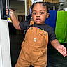 toddler, child, overalls, black_shirt, door_handle, indoor, kitchen, curly_hair, pigtails, young_child, standing, expression, person, floor, cabinet, refrigerator, bag, colorful_background, home, curiosity