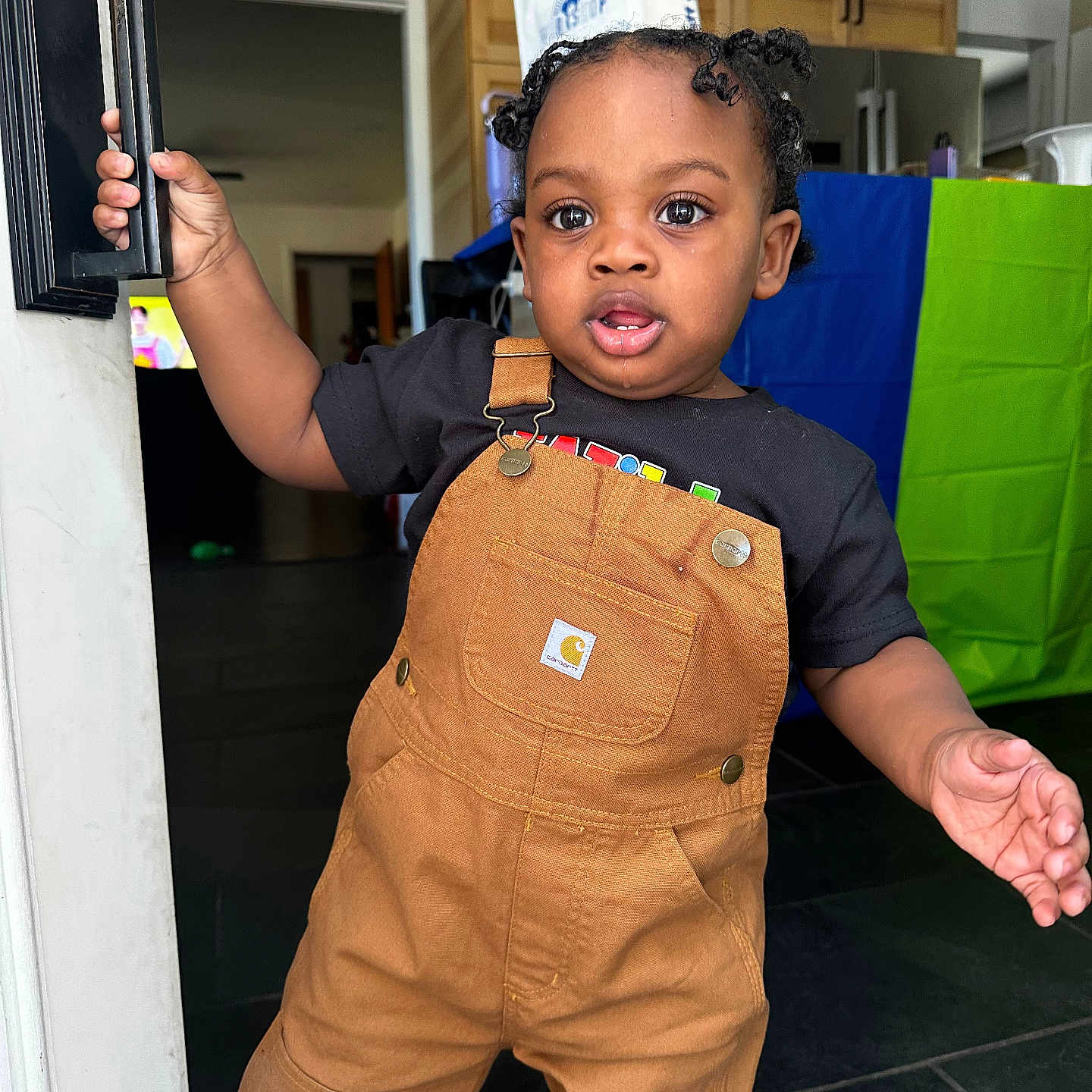 Yosiah joined the competition — help win amazing prizes! toddler, child, overalls, black_shirt, door_handle, indoor, kitchen, curly_hair, pigtails, young_child, standing, expression, person, floor, cabinet, refrigerator, bag, colorful_background, home, curiosity