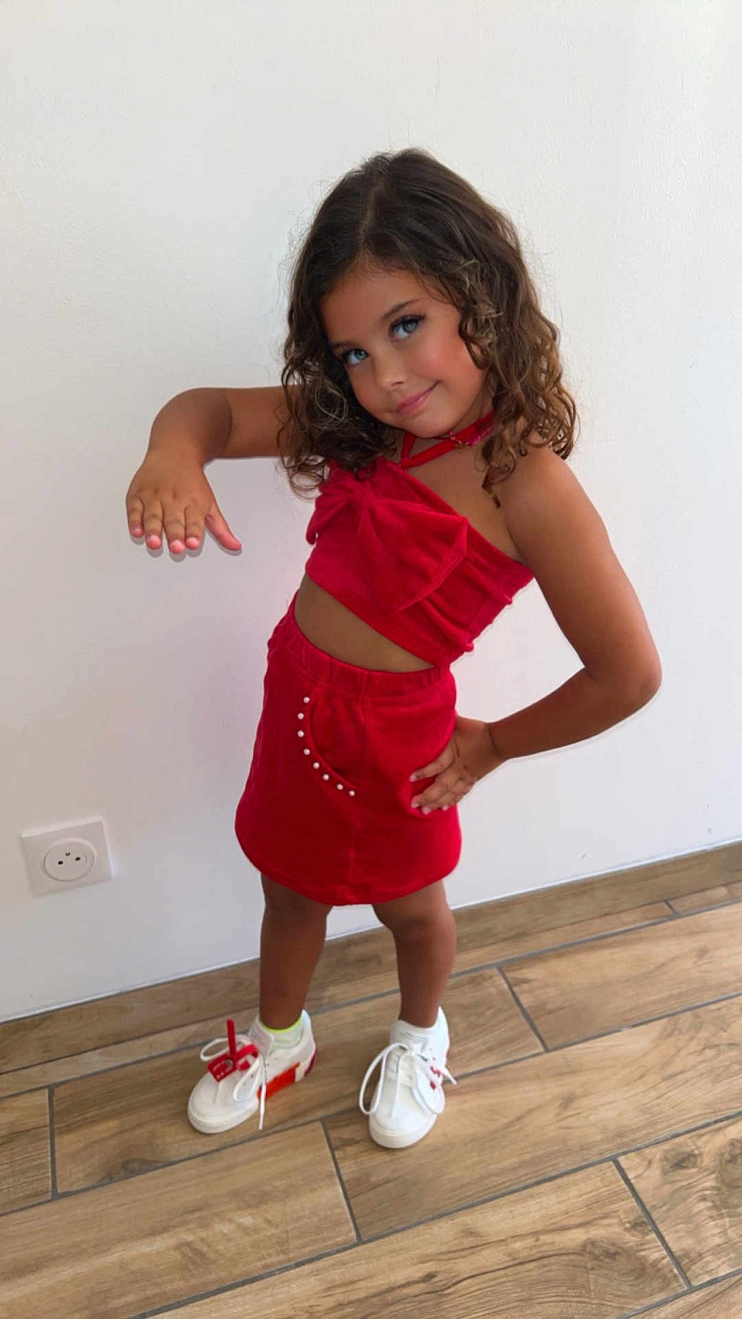 Hatlanna participe au concours pour gagner de l'argent avec cette photo : child, girl, red_clothing, curly_hair, smile, pose, white_wall, wooden_floor, sneakers, fashion, playful, cute, indoor, happy, portrait, standing, mischievous, casual, modern, accessories