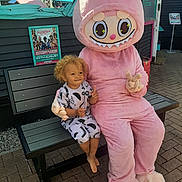 Mateus is registered to the contest to win money with this photo: child, toddler, bench, mascot, costume, pink, outdoor, sunny, smiling, curly_hair, barefoot, peace_sign, pavement, building, poster, event, daytime, casual_clothing, happy, person