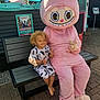 child, toddler, bench, mascot, costume, pink, outdoor, sunny, smiling, curly_hair, barefoot, peace_sign, pavement, building, poster, event, daytime, casual_clothing, happy, person