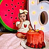 Eliana joined the competition — help win amazing prizes! baby, birthday, bow, cake, celebration, child, dessert, food, fruit, hair_bow, indoor, marquee_lights, party, pink_blanket, portrait, red_shorts, sitting, smiling, strawberry, watermelon