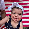 Eliana joined the competition — help win amazing prizes! baby, camera_facing, child, close_up, dark_clothing, face, happy, headwrap, infant, joyful, outdoor, person, photography, portrait, red_and_white, seated, smiling, star_pattern, striped_clothing, white_headwrap
