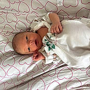 Freya is registered to the contest to win money with this photo: newborn, baby, infant, lying_down, onesie, white_clothing, heart_pattern, bed_sheet, cute, small_hands, face, eyes_open, fabric, soft, sleeping_area, indoor, child, person, resting, cozy