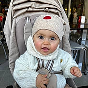 Luisa participe au concours pour gagner de l'argent avec cette photo : baby, child, stroller, hat, knit_sweater, indoor, seat_belt, curious, cute, infant, face, hand, clothing, person, seated, cafe, table, chair, brown_pants, expression