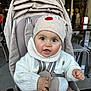 baby, child, stroller, hat, knit_sweater, indoor, seat_belt, curious, cute, infant, face, hand, clothing, person, seated, cafe, table, chair, brown_pants, expression