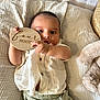 baby, infant, child, four_months, holding_sign, white_shirt, green_pants, plush_toy, blanket, soft_texture, curious_eyes, lying_down, wooden_sign, indoor, portrait, cute, small_hands, buttoned_shirt, baby_clothes, resting