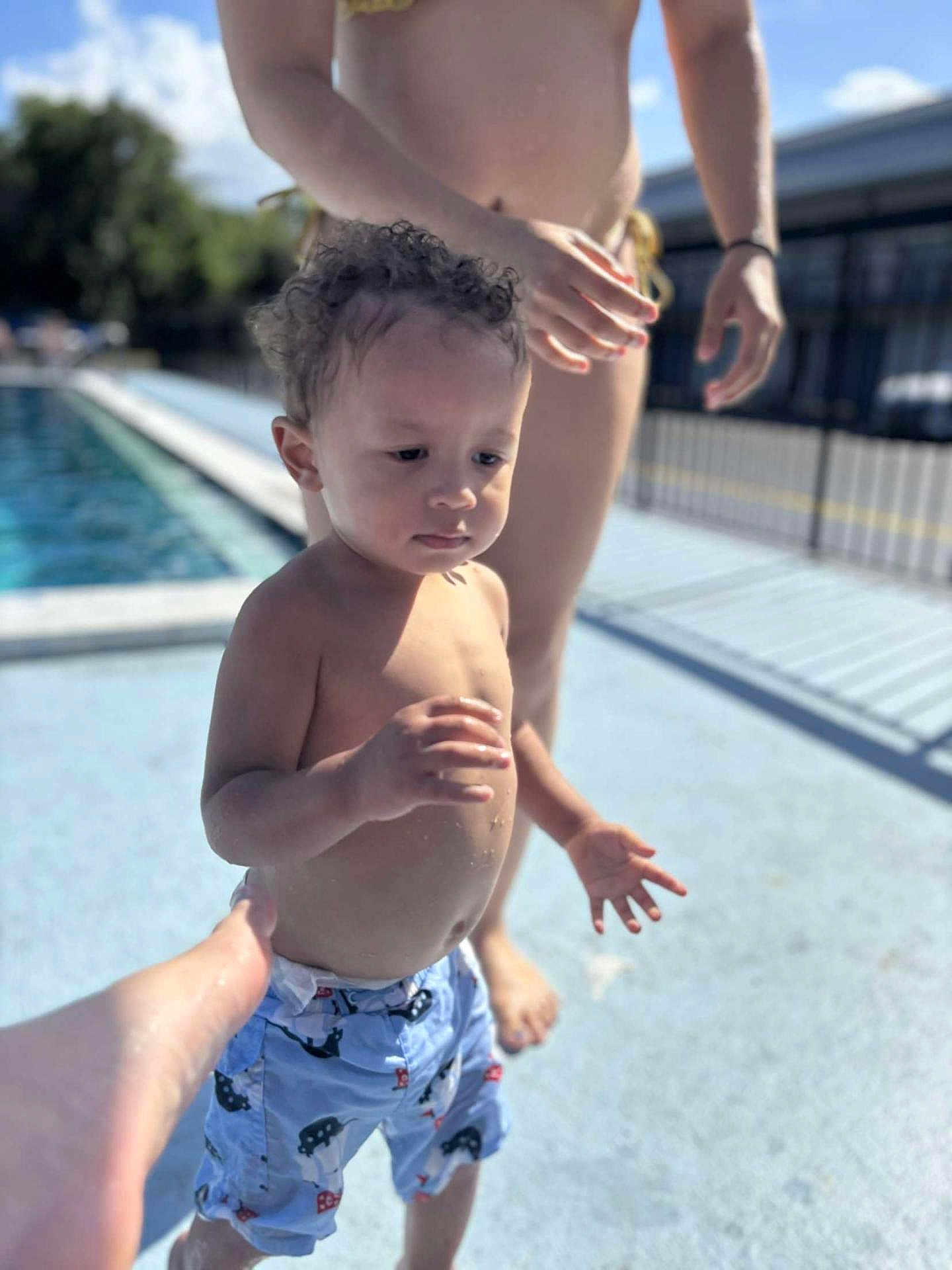Weston joined the competition — help win amazing prizes! toddler, child, poolside, swim_trunks, water, adult, hand, sunlight, outdoor, summer, curly_hair, barefoot, blue_sky, fence, pavement, person, standing, protective, daytime, casual