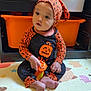 baby, child, halloween_outfit, pumpkin_design, orange_clothing, play_mat, toy, sitting, indoors, storage_bin, shelf, plastic_toy, headwear, cute, infant, footwearless, expression, face, person, playroom